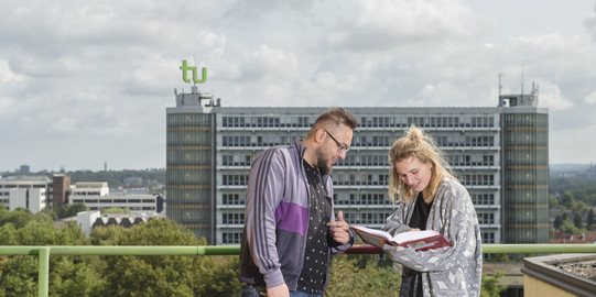 A male and a female student are looking into a book together, in the background the Math Tower can be seen in its full length.