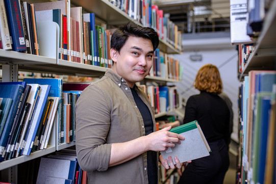 Student stands in front of a bookshelf with a book in his hands and smiles in the camera.